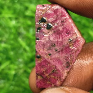 Peut inclure: Une pierre précieuse brute et allongée d'une teinte magenta et rose profond, présentant une surface cristalline texturée. La pierre affiche des inclusions naturelles visibles, dont de petites taches noires éparses et des veines minérales brun doré.