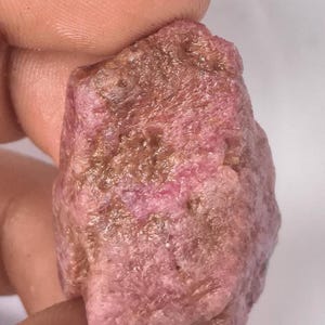 May include: A raw, unpolished gemstone with a rough, textured surface. The stone displays a mix of deep pink and earthy brown tones throughout its irregular, blocky shape, showcasing natural mineral formations and a matte, opaque appearance.