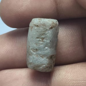 Puede incluir: Una pequeña piedra preciosa en bruto de forma cilíndrica, con un color blanquecino a gris claro translúcido. La superficie de la piedra es irregular y texturizada, con sutiles inclusiones marrones y hendiduras naturales en toda su estructura.
