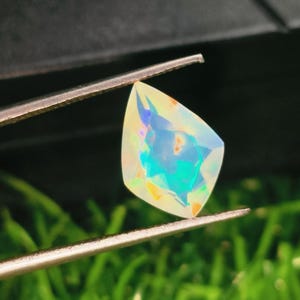 May include: A faceted, pear-shaped opal gemstone held by metal tweezers. The stone displays a translucent, milky white base colour with vibrant flashes of iridescent blue, green, and orange light reflecting from within its polished, multi-faceted surface.