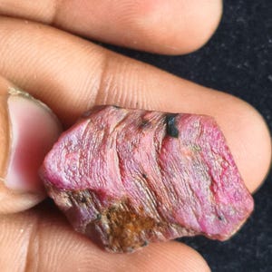 Könnte beinhalten: Ein roher, unpolierter Rubin-Edelstein mit einem leuchtenden rosa-roten Farbton. Der Stein weist eine ausgeprägte, geschichtete und gestreifte Oberflächenstruktur mit sichtbaren natürlichen Einschlüssen und dunklen Mineralablagerungen auf.