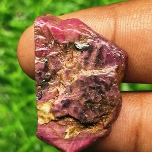 Op de afbeelding: Een ruwe, ongepolijste robijn met een diepe magenta en karmozijnrode tint. De steen heeft een ruw, getextureerd oppervlak met zichtbare natuurlijke insluitingen, donkere minerale aders en vlekken van wit kristallijn materiaal.
