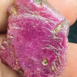 May include: A raw, uncut ruby gemstone with a vibrant magenta-pink hue. The stone features a natural, irregular shape with a textured, crystalline surface, visible internal fractures, and small inclusions of metallic-colored mineral deposits.
