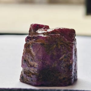 May include: A raw, uncut ruby gemstone with a deep magenta and purple hue. The stone features a rough, crystalline texture with visible dark brown and black mineral inclusions embedded within its faceted, irregular surface structure.