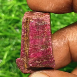 Puede incluir: Una piedra preciosa de rubí en bruto y sin tallar de color magenta y carmesí intenso. La piedra presenta una superficie rugosa y texturizada con estrías naturales visibles e inclusiones de color marrón terroso en un lado.