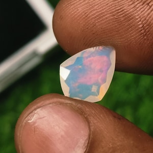 May include: A pear-shaped opal gemstone featuring a translucent white base with iridescent flashes of pink, blue, and orange light. The stone is faceted with a geometric pattern that reflects light across its surface, displaying a play of color.