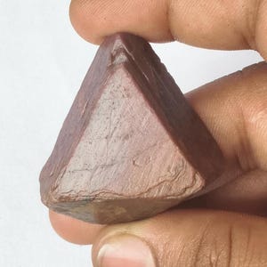 May include: A raw, triangular-shaped gemstone with a deep reddish-brown hue and a textured, matte surface. The stone features visible natural striations and irregular edges, showcasing a solid, opaque appearance with earthy, muted tones throughout.