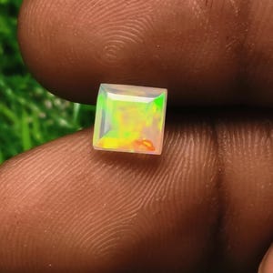 May include: A square-cut opal gemstone featuring a translucent body with vibrant flashes of green, yellow, and orange play-of-color. The gemstone has a faceted surface that reflects light, showcasing a multi-colored iridescent appearance throughout.