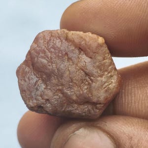 May include: A raw, unpolished gemstone with a hexagonal crystal structure and a brownish-pink hue. The surface of the stone displays a natural, textured appearance with visible striations and a semi-translucent quality throughout the mineral.