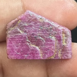Puede incluir: Una piedra preciosa de rubí en bruto y sin pulir con un tono magenta rosado intenso. La piedra presenta una base rectangular plana con un borde superior triangular puntiagudo. Su superficie está texturizada con inclusiones naturales.