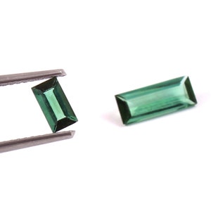 May include: Two rectangular, faceted green gemstones with a baguette cut. One gemstone is held by metal tweezers, while the other is positioned to the right. The stones exhibit a deep, translucent emerald green hue with sharp edges.