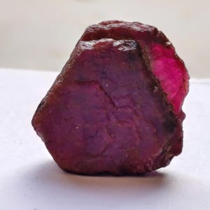 Peut inclure: Une pierre précieuse rubis brute et non polie d'une couleur magenta profonde et rouge foncé. La pierre présente une surface rugueuse et texturée avec des facettes naturelles et des bords irréguliers, révélant un éclat rose vif translucide.
