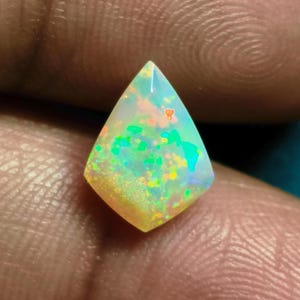 May include: A kite-shaped opal gemstone featuring a translucent white base with vibrant flashes of green, orange, and yellow play-of-color throughout its faceted surface. The stone displays a polished finish with sharp, angular edges and a pointed top.
