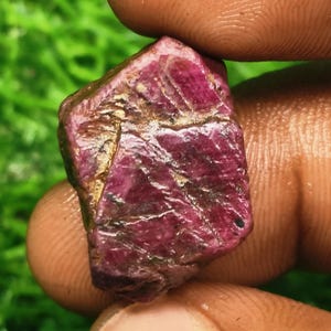 Peut inclure: Une pierre précieuse brute et non taillée d'une couleur magenta profond et rouge foncé. La pierre présente une surface rugueuse et irrégulière avec des fissures naturelles visibles, des textures cristallines et des sections opaques.