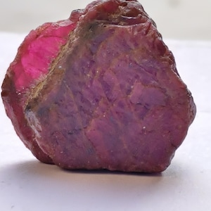 Puede incluir: Una gema de rubí en bruto, de color rojo intenso y púrpura. La piedra natural tiene una superficie texturizada y diferentes tonos, con una zona rosa brillante en un lado. Esta gema en bruto es un mineral natural.