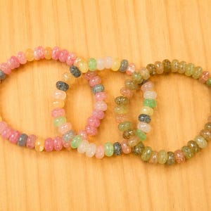 May include: Three circular bracelets composed of small, translucent, disc-shaped beads. The beads feature a variety of colours including shades of pink, yellow, green, and grey, arranged in a repeating pattern along each elasticised strand.