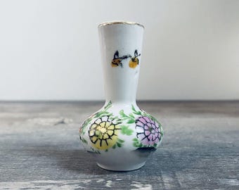 May include: A small white ceramic vase with a floral design. The vase has a narrow neck and a round body. The flowers are painted in shades of pink, yellow, and green.