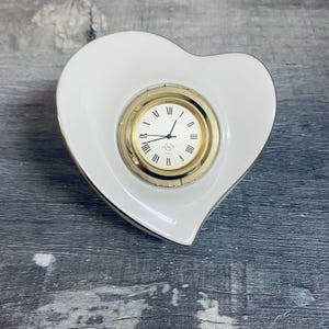 May include: A white heart-shaped clock with a gold-tone clock face and Roman numerals. The clock has a gold trim and the brand name "Lenox" is visible on the clock face. The clock is on a gray wooden surface.