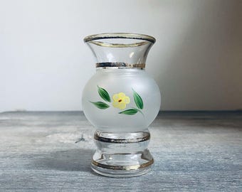 May include: A small, frosted glass vase with a round body and a clear, flared top. The vase features a hand-painted yellow flower with green leaves. Gold trim accents the top and base, adding a touch of elegance to this decorative piece.