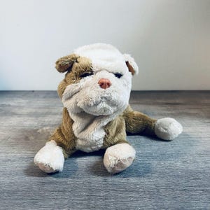 May include: A plush bulldog toy featuring soft, textured fur in shades of brown and white. The stuffed animal has a wrinkled face, a small pink nose, and floppy ears, depicted in a lying-down position with its front paws extended forward.
