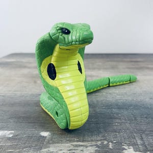 May include: A green and yellow plastic cobra toy with black eye details. The toy snake is posed in a striking, upright position, with its hood expanded. The segmented body extends to the right, suggesting movement. The toy is on a gray wooden surface.