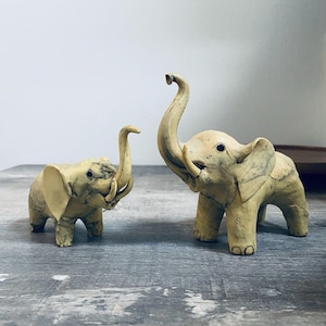 May include: Two small, off-white elephant figurines with raised trunks and visible tusks. The elephants have detailed features, including ears and eyes, and are positioned on a gray surface.