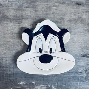 May include: A white ceramic dish shaped like a cartoon cat face, featuring black and white details. The dish has large eyes, a black nose, and a smiling mouth. The dish is on a gray wooden surface.