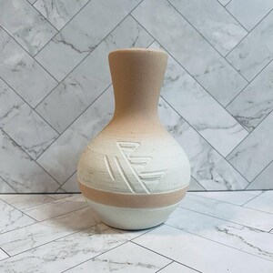 May include: A ceramic vase with a narrow neck and a rounded body, featuring a two-tone design of matte beige and white. The surface displays a carved geometric pattern consisting of angled lines etched into the white section of the vessel.