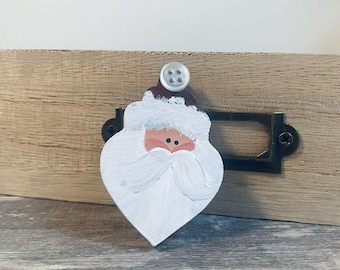 May include: A hand-painted wooden Santa Claus ornament. The ornament features a white beard, a red nose, and a brown hat with white trim. A white button is attached to the top. The ornament is placed on a wooden surface.