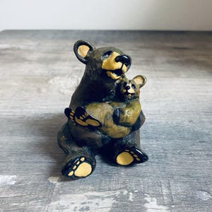 May include: A brown bear figurine holding a smaller bear cub. The bears have yellow accents on their faces and paws. The figurine is sitting on a gray surface.