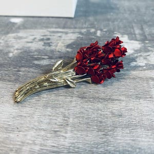 May include: A gold-toned metal brooch in the shape of a bouquet of red flowers. The flowers are clustered together, with detailed leaves and stems. The brooch is likely a vintage or costume jewelry piece.