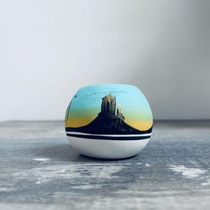 May include: A small, round, hand-painted ceramic pot with a landscape design. The pot features a blue sky, a black mountain, and a yellow and orange sunset. The bottom half is white with a black stripe. The pot is sitting on a gray wooden surface.