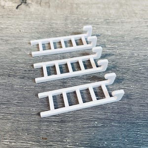 May include: Three small, white, plastic ladders. Each ladder has a series of horizontal rungs and a curved hook at the top. The ladders are arranged diagonally on a gray, textured surface.