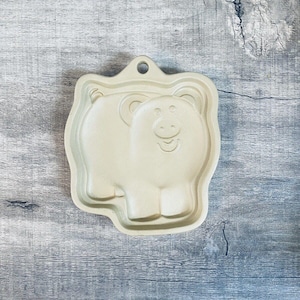 May include: A light beige, pig-shaped baking mold with a smiling face and a loop for hanging. The mold has a rectangular shape with rounded edges and is made of a solid material. The pig's features are subtly embossed.