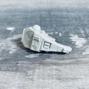 May include: A light gray, plastic model of a spaceship component. The piece has a rounded top with a small dome, a rectangular body with detailed panels, and a spiral design on one end. The object is on a gray surface.