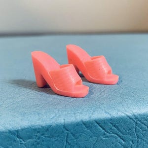 May include: A pair of pink, open-toe high-heeled sandals. The heels are thick and the sandals have a textured surface. The shoes are made of a solid, opaque material.