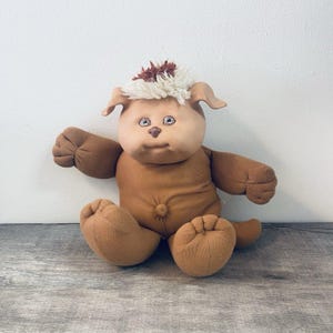 May include: A plush doll with a tan fabric body, a round head, and a brown nose. The doll features a tuft of white and brown yarn hair on top of its head, floppy ears, and light-colored eyes. It has a soft, stuffed torso with a navel detail.