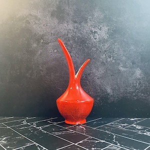 May include: A vibrant red ceramic vase with a unique, elongated shape. The vase features a bulbous base and two slender, upward-curving spouts. The interior is white, contrasting with the speckled red exterior. The vase is a decorative item.