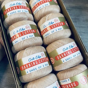 May include: A collection of beige darning cotton balls arranged in a cardboard box. Each ball features a paper label with the text Gilt Edge DARNING COTTON 4 ENDS OF 2 PLY and a small circular icon with a star and the number 10 inside a red circle.