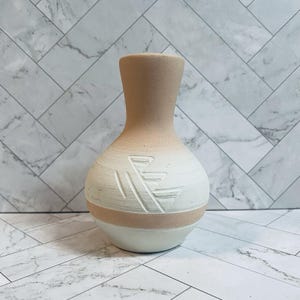 May include: A ceramic vase with a rounded body and a narrow neck, featuring a two-tone color scheme of matte beige and off-white. The surface displays a carved geometric pattern consisting of angled lines on the front of the rounded section.