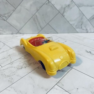 May include: A yellow toy convertible car with a red interior. The car has a clear windshield and black wheels. The car's design is reminiscent of classic sports cars.