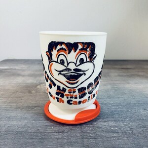 May include: A white plastic cup featuring a black and orange graphic of a cartoon character with a mustache. The text reads South of the Border South Carolina. The cup has a white base and a detachable orange plastic ring at the bottom.