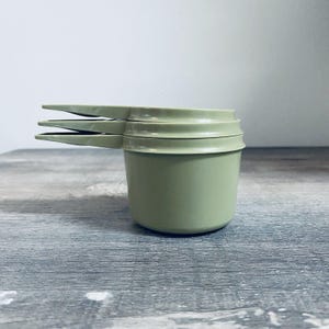 May include: A stack of three sage green measuring cups. Each cup has a rounded base and a long, flat handle extending from the rim. The cups are made of a matte material and are stacked neatly on top of each other.