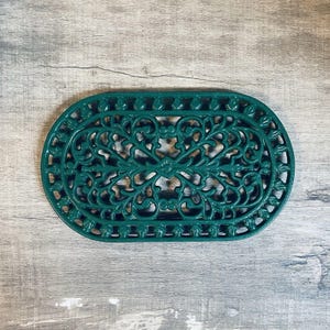 May include: An oval-shaped dark green trivet featuring an intricate, openwork scroll and leaf pattern. The metal object has a symmetrical design with a decorative border consisting of repeating leaf-like motifs around the entire outer edge.