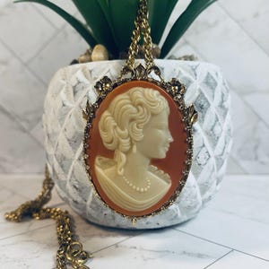 May include: A vintage cameo pendant necklace. The oval pendant features a cream-colored profile of a woman set against an orange background. The pendant is framed in gold-tone metal with floral accents and hangs from a gold chain.