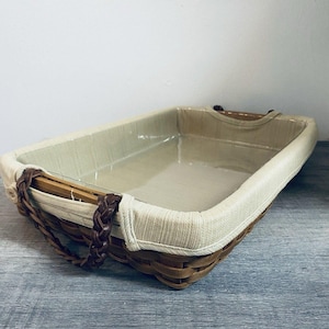 May include: Rectangular woven basket with a light-colored fabric lining. The basket has a brown woven base and sides, with a braided handle on each end. The interior is lined with a beige fabric. The basket is empty.