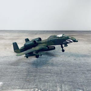 May include: A green toy airplane with a white and black shark mouth design on the nose. The toy plane has two engines, wings, and a tail. The toy is made of plastic and is sitting on a wooden surface.
