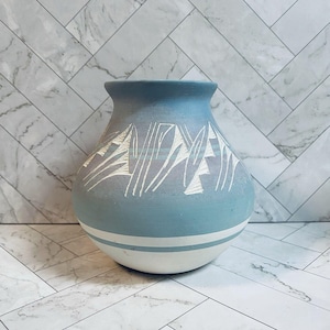 May include: A matte light blue vase with a wide, rounded body and a flared rim. The exterior features white etched geometric patterns around the upper section and two horizontal white stripes encircling the lower portion of the vessel.