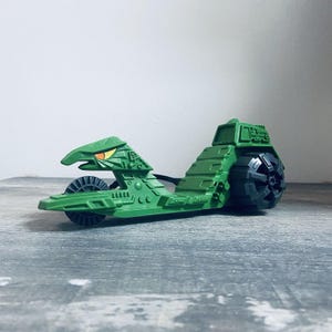 May include: A green toy vehicle with a snake-like head and a large wheel on the back. The vehicle has a detailed design with various shapes and textures. The head has yellow eyes. The toy is on a gray surface.