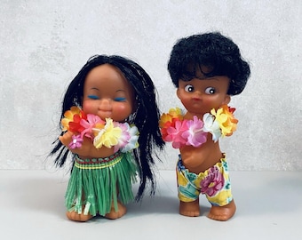 May include: Two small dolls with dark skin and black hair. The left doll has long hair, a green grass-style skirt, and a colorful floral lei. The right doll has short curly hair, yellow floral shorts, and a matching colorful floral lei around its neck.